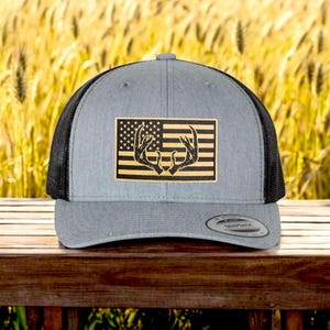 Pode incluir: Um chapéu de caminhoneiro cinza e preto com um remendo de uma bandeira americana com um par de chifres de veado no meio.