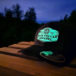Könnte beinhalten: Eine schwarze Baseballkappe mit einem im Dunkeln leuchtenden Patch, auf dem in weißen Buchstaben "EST. 2020 BLUE COLLAR APPAREL" steht. Der Patch hat eine Holzmaserung und eine kleine amerikanische Flagge in der oberen linken Ecke.
