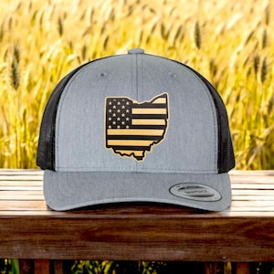 May include: A gray and black trucker hat with a gold outline of the state of Ohio with an American flag design inside. The hat is sitting on a wooden surface.