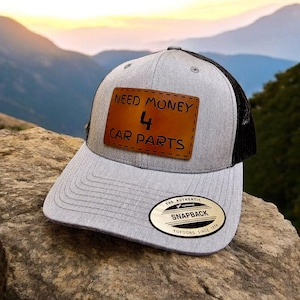 Puede incluir: Una gorra de camionero gris y negra con un parche de cuero marrón que dice "Need Money 4 Car Parts". La gorra tiene una malla negra en la parte posterior y una etiqueta de cuero marrón que dice "The Authentic Snapback Yupoong Since 1974".