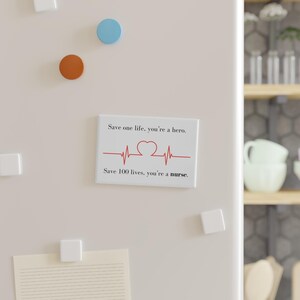 Op de afbeelding: Witte rechthoekige magneet met de tekst "Save one life, you're a hero. Save 100 lives, you're a nurse." in zwarte letters. Een rode hartslaglijn met een hartvorm staat in het midden.