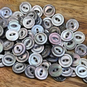 Puede incluir: Un conjunto de botones iridiscentes y nacarados, cada uno con dos agujeros. Los botones son en tonos grises, blancos y negros, y están dispersos sobre una superficie de madera. Ideales para costura y manualidades.