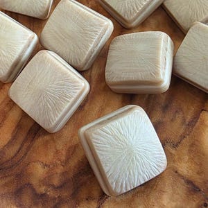 May include: A collection of eleven square, off-white buttons. Each button features a raised, beveled edge and a textured surface with a radiating, sunburst-like pattern that originates from the center of each individual piece.