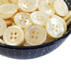 May include: A bowl filled with white pearl buttons with four holes each. The buttons are smooth and shiny.