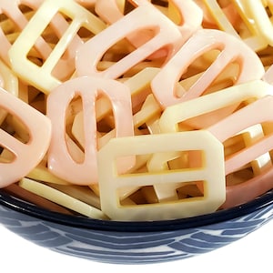 May include: A bowl filled with a variety of plastic buckles in two colors: pale pink and cream. The buckles are all the same shape, with a rectangular body and two oval holes in the center.
