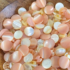 May include: A collection of round, smooth buttons in shades of peach, soft pink, and iridescent white. Each button features a polished, reflective surface with a subtle luster, characteristic of shell material, and includes two small holes for attachment.