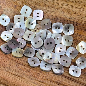 May include: A selection of square mother-of-pearl buttons, each with two holes, in shades of white, grey, and brown. The buttons are scattered on a wooden surface. Suitable for sewing and craft projects, these buttons offer a classic aesthetic.