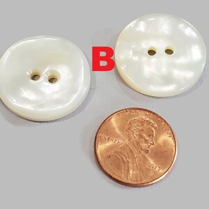 May include: Two large, round, off-white buttons with two holes each. A penny is next to the buttons for size comparison. The buttons have a pearlescent sheen, suggesting a mother-of-pearl material. The buttons are on a light gray surface.