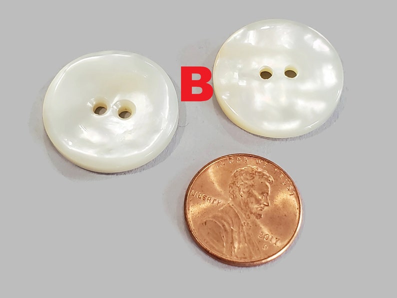 May include: Two large, round, off-white buttons with two holes each. A penny is next to the buttons for size comparison. The buttons have a pearlescent sheen, suggesting a mother-of-pearl material. The buttons are on a light gray surface.