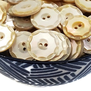 May include: A bowl filled with a variety of white and cream colored mother of pearl buttons. The buttons have two holes and a decorative edge.
