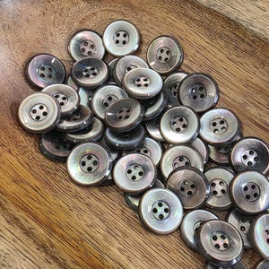 May include: A collection of round, four-hole buttons with a smooth, iridescent finish. The buttons feature a dark, pearlescent gray color with subtle variations in tone, creating a reflective surface. Each button has four centered holes.