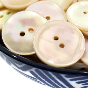May include: A bowl of white iridescent buttons with two holes each. The buttons are smooth and shiny, and they are arranged in a random pattern.