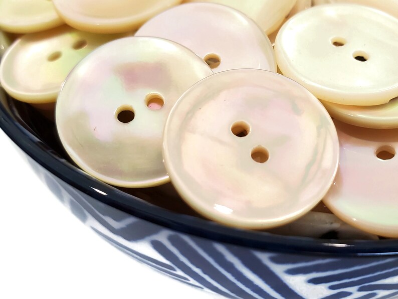 May include: A bowl of white iridescent buttons with two holes each. The buttons are smooth and shiny, and they are arranged in a random pattern.