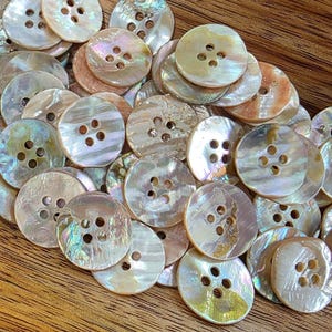 May include: A pile of iridescent, round buttons, each with four holes. The buttons have a pearlescent sheen, with colours from white and cream to hints of pink and green. They are scattered on a wooden surface.