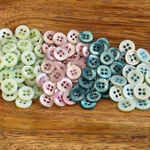 May include: A collection of small, round, four-hole buttons in three distinct color groups: light mint green, soft pastel pink, and deep teal blue. Each button features a smooth, slightly iridescent finish and a recessed center area for thread.
