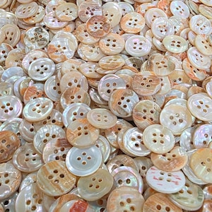 May include: A large collection of round, four-hole buttons made from iridescent mother-of-pearl. The buttons feature a variety of natural cream, beige, and light brown marbled patterns with a smooth, glossy finish and a slightly raised center.