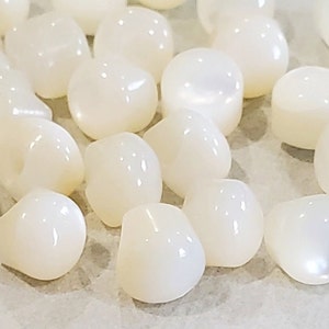 May include: A collection of white, pearlescent, round, and slightly oval-shaped beads. The beads have a smooth, glossy finish.