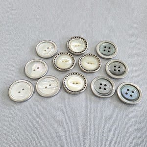 May include: A collection of round buttons in various designs. Some buttons have a pearlescent center with two or four holes, while others have a decorative silver-colored border. The buttons are arranged on a light gray surface.