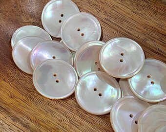 May include: A collection of iridescent, round buttons with two holes each. The buttons have a pearlescent sheen and are scattered on a wooden surface. These buttons are ideal for sewing and craft projects.