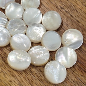 May include: A collection of round, white mother-of-pearl buttons with a smooth, iridescent finish. Each button features a slightly convex surface with natural variations in luster and texture, displaying a soft, pearlescent sheen across the surface.