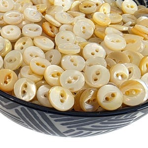 May include: A close-up shot of a bowl filled with numerous round, off-white buttons. The buttons have two holes and a slightly iridescent sheen. Some buttons have a golden hue. The bowl has a black and white patterned rim.