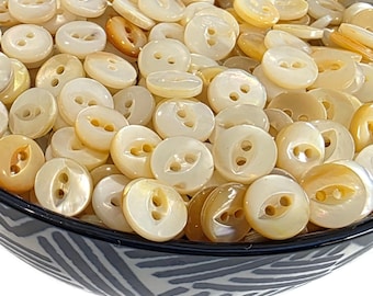 May include: A close-up shot of a bowl filled with numerous round, off-white buttons. The buttons have two holes and a slightly iridescent sheen. Some buttons have a golden hue. The bowl has a black and white patterned rim.