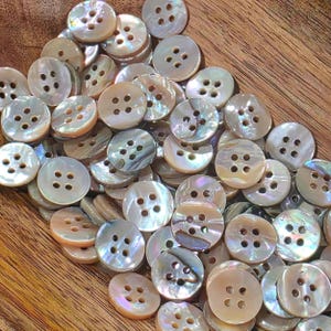 May include: A collection of round, four-hole buttons crafted from iridescent mother-of-pearl shell. Each button features a smooth, polished surface with natural variations in color, displaying subtle shades of cream, beige, and soft pearlescent tones.