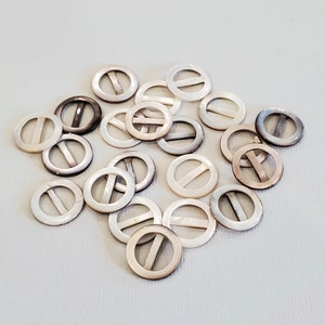 May include: A collection of round, mother-of-pearl belt buckles with a single bar across the center. The buckles vary in color from white to dark grey, and are scattered on a light background.