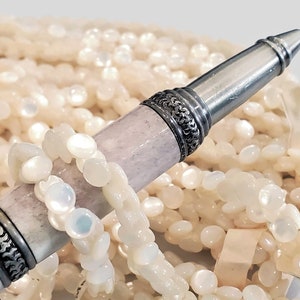 May include: A silver pen with a white and grey patterned barrel, lying on a bed of white, iridescent, round beads.