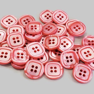Può includere: Un mucchio di bottoni di plastica rosa a quattro fori. I bottoni sono tutti della stessa dimensione e forma, e hanno una finitura leggermente iridescente.