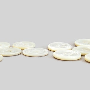 May include: A collection of off-white, round buttons with a pearlescent sheen. Each button has a central hole and a slightly raised rim. The buttons are arranged on a light gray surface, showcasing their subtle color variations.