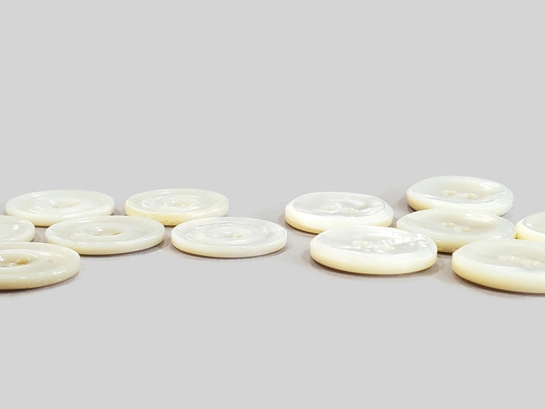 May include: A collection of off-white, round buttons with a pearlescent sheen. Each button has a central hole and a slightly raised rim. The buttons are arranged on a light gray surface, showcasing their subtle color variations.