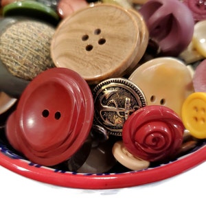 May include: A bowl filled with a variety of colorful buttons, including red, brown, gold, white, and yellow. The buttons are made of different materials, such as plastic, metal, and wood.