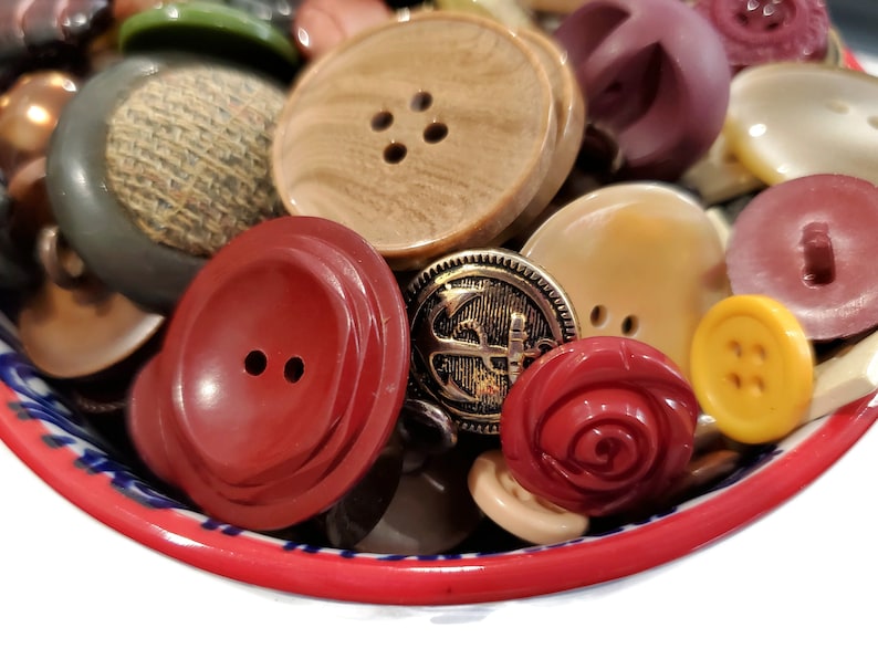 May include: A bowl filled with a variety of colorful buttons, including red, brown, gold, white, and yellow. The buttons are made of different materials, such as plastic, metal, and wood.