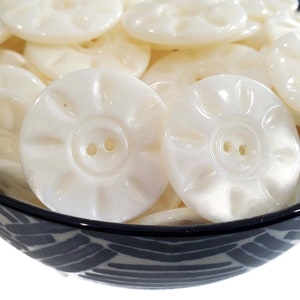 May include: A bowl filled with white plastic buttons with a raised flower design and two holes. The buttons are in a variety of sizes and are all white.
