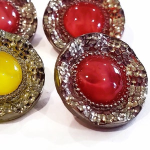 May include: Close-up of several decorative buttons. The buttons have a metallic, textured border and a central, glossy, round gem. The gems are in shades of red and yellow. The buttons are on a white background.
