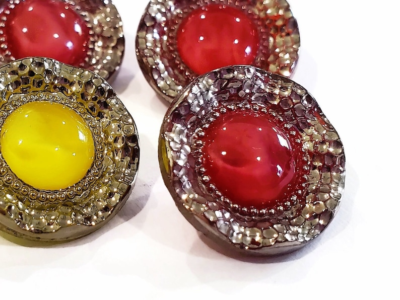 May include: Close-up of several decorative buttons. The buttons have a metallic, textured border and a central, glossy, round gem. The gems are in shades of red and yellow. The buttons are on a white background.