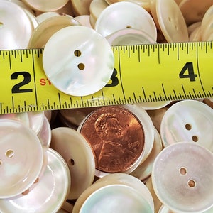 May include: A close-up of a pile of white iridescent buttons with two holes each. A measuring tape is placed on top of the buttons, showing that they are approximately 1 inch in diameter.