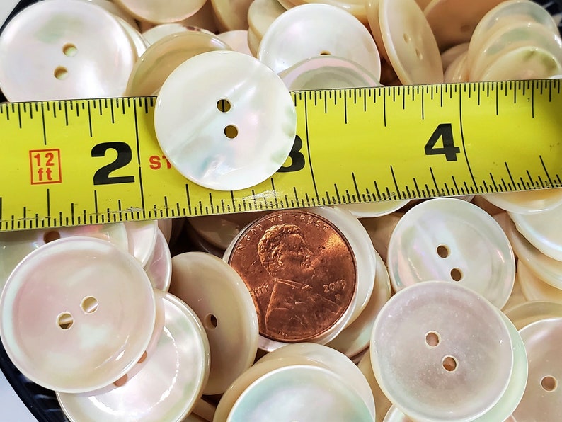 May include: A close-up of a pile of white iridescent buttons with two holes each. A measuring tape is placed on top of the buttons, showing that they are approximately 1 inch in diameter.