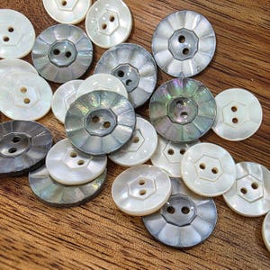 May include: A collection of iridescent and white shell buttons. The buttons have two holes and a variety of designs, including a scalloped edge and a hexagonal shape. The buttons are scattered on a wooden surface.