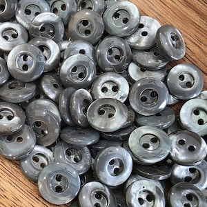 Puede incluir: Una colección de botones redondos de dos agujeros con un acabado gris oscuro brillante. Cada botón presenta un centro liso y ligeramente cóncavo con dos orificios circulares para su fijación, mostrando un sutil brillo jaspeado.