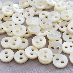 Puede incluir: Un conjunto de pequeños botones redondos, de color blanco roto, con dos agujeros cada uno. Los botones están dispersos, mostrando su acabado liso y ligeramente iridiscente. Ideal para costura y manualidades.