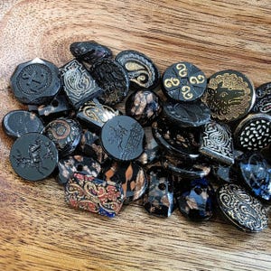 Könnte beinhalten: Eine Sammlung schwarzer Vintage-Knöpfe mit verschiedenen Designs, darunter Paisley-, geometrische und Tiermotive. Einige Knöpfe weisen goldene oder kupferfarbene Akzente auf. Die Knöpfe sind auf einer Holzoberfläche angeordnet.