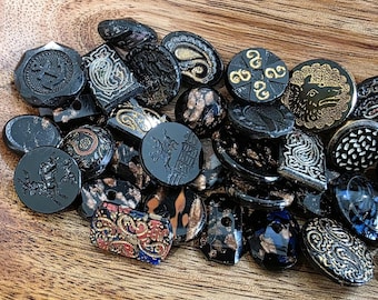 May include: A collection of vintage black buttons with various designs, including paisley, geometric patterns, and animal motifs. Some buttons feature gold or copper accents. The buttons are arranged on a wooden surface.