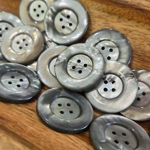 May include: A collection of iridescent, round buttons with four holes each. The buttons have a pearlescent sheen, with shades of gray, white, and cream. The buttons are arranged on a wooden surface.