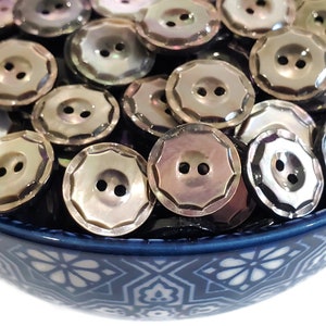 May include: A bowl filled with iridescent, two-hole, mother-of-pearl buttons. The buttons have a scalloped edge and a shiny, pearlescent finish.