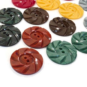 May include: A collection of round, two-hole buttons in various colors including dark green, brown, rust, yellow, and gray. Each button features a distinct swirling, radial pattern carved into the surface, creating a textured, fan-like appearance.