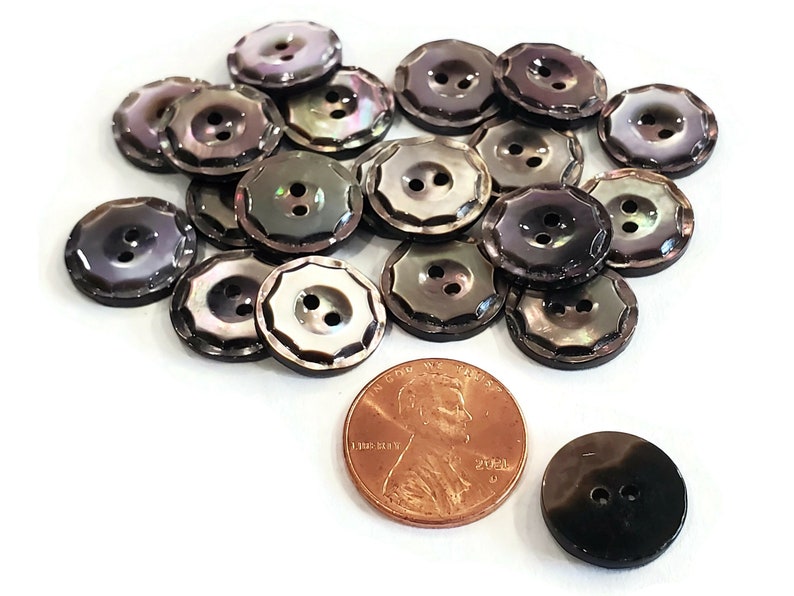 May include: A pile of 18 black and iridescent shell buttons with two holes each. The buttons have a scalloped edge and are approximately the size of a US penny.