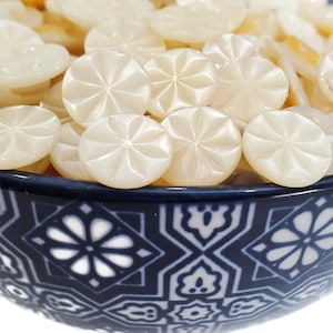 May include: A bowl filled with white, round, decorative buttons with a carved starburst design. The buttons are in a blue and white bowl with a floral pattern.