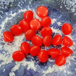 May include: A collection of bright red, glossy, oval-shaped candies scattered on a blue and white patterned surface. The candies have a smooth texture and are evenly distributed across the surface.